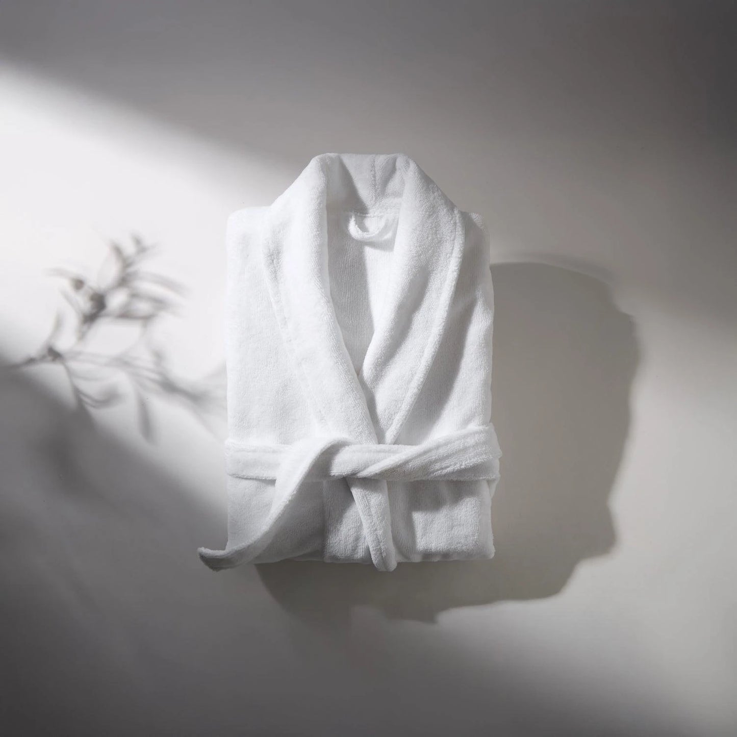 White bathrobe made of soft fabric, placed on a light surface with soft shadow, creating an atmosphere of comfort and relaxation.