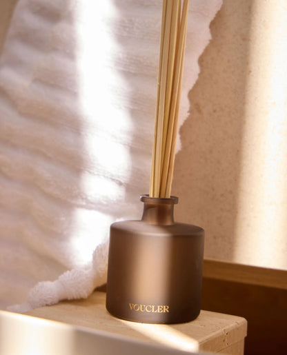 Voucler brown aroma diffuser placed on a wooden table, next to a white towel and decorated with a stone wall and beige wall in the background.
