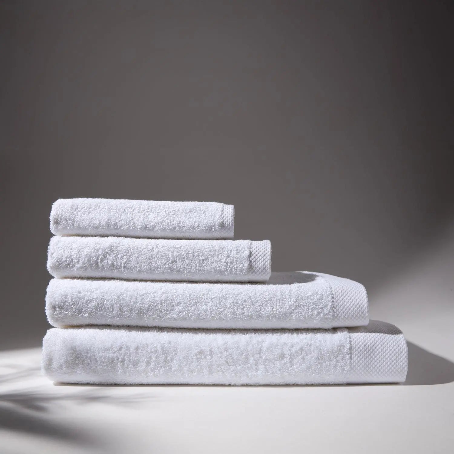 Image of four white towels stacked on top of each other, with a soft gray background, conveying a feeling of cleanliness and care for hygiene.
