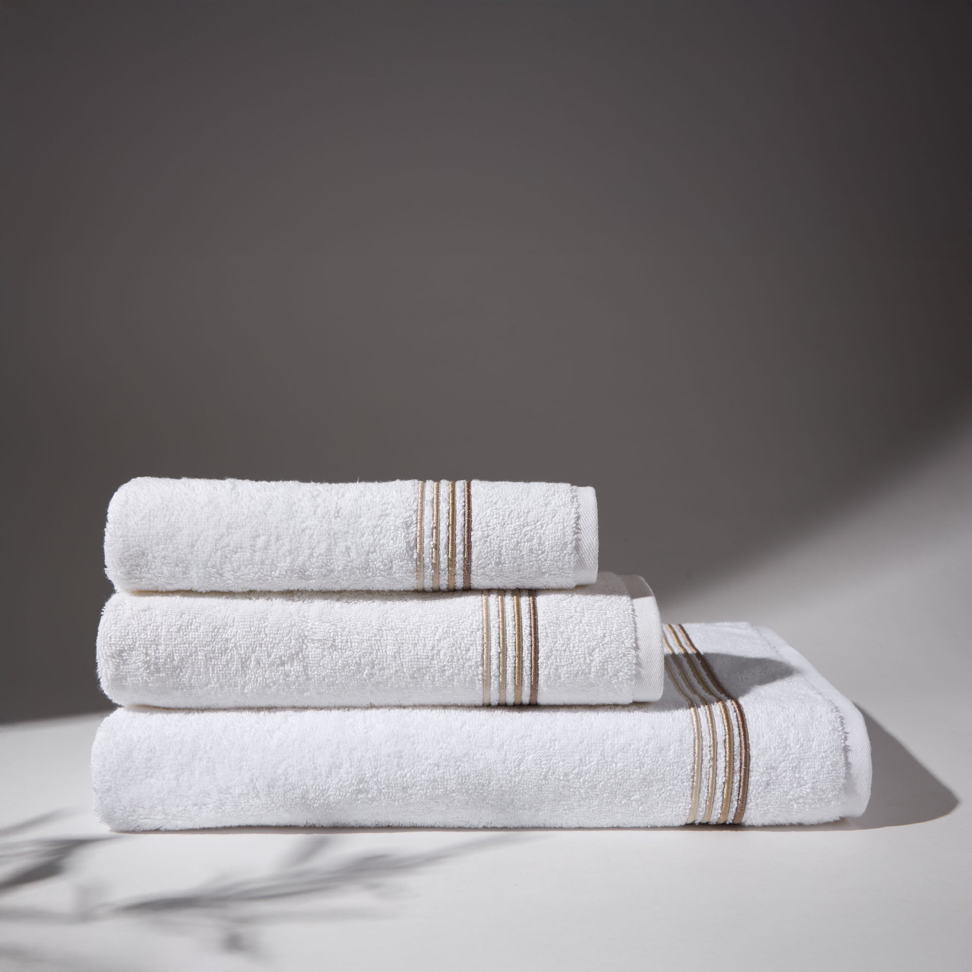 Set of white towels with beige stripes stacked under soft lighting, on a flat surface and neutral background.