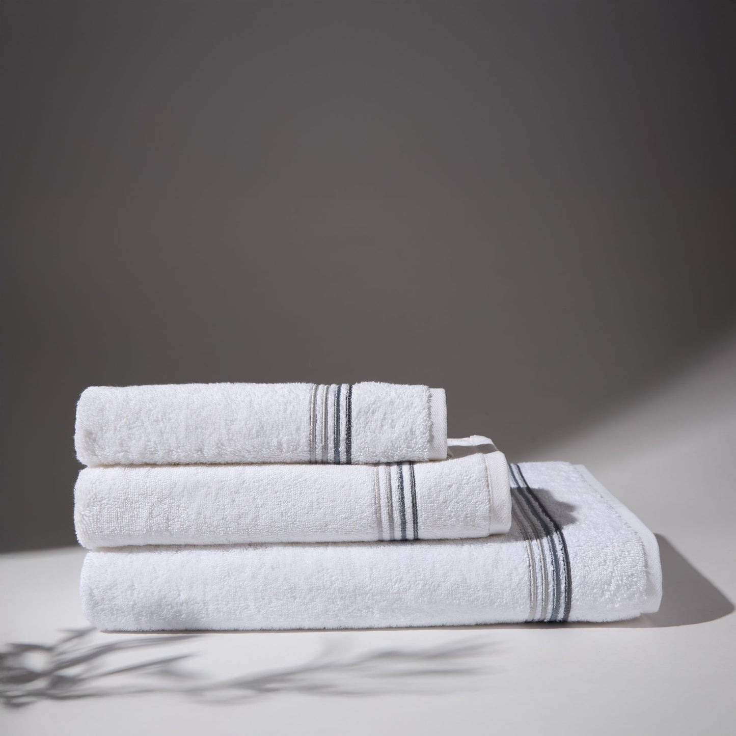 Set of three white towels with gray stripes folded on a surface with a gray background.