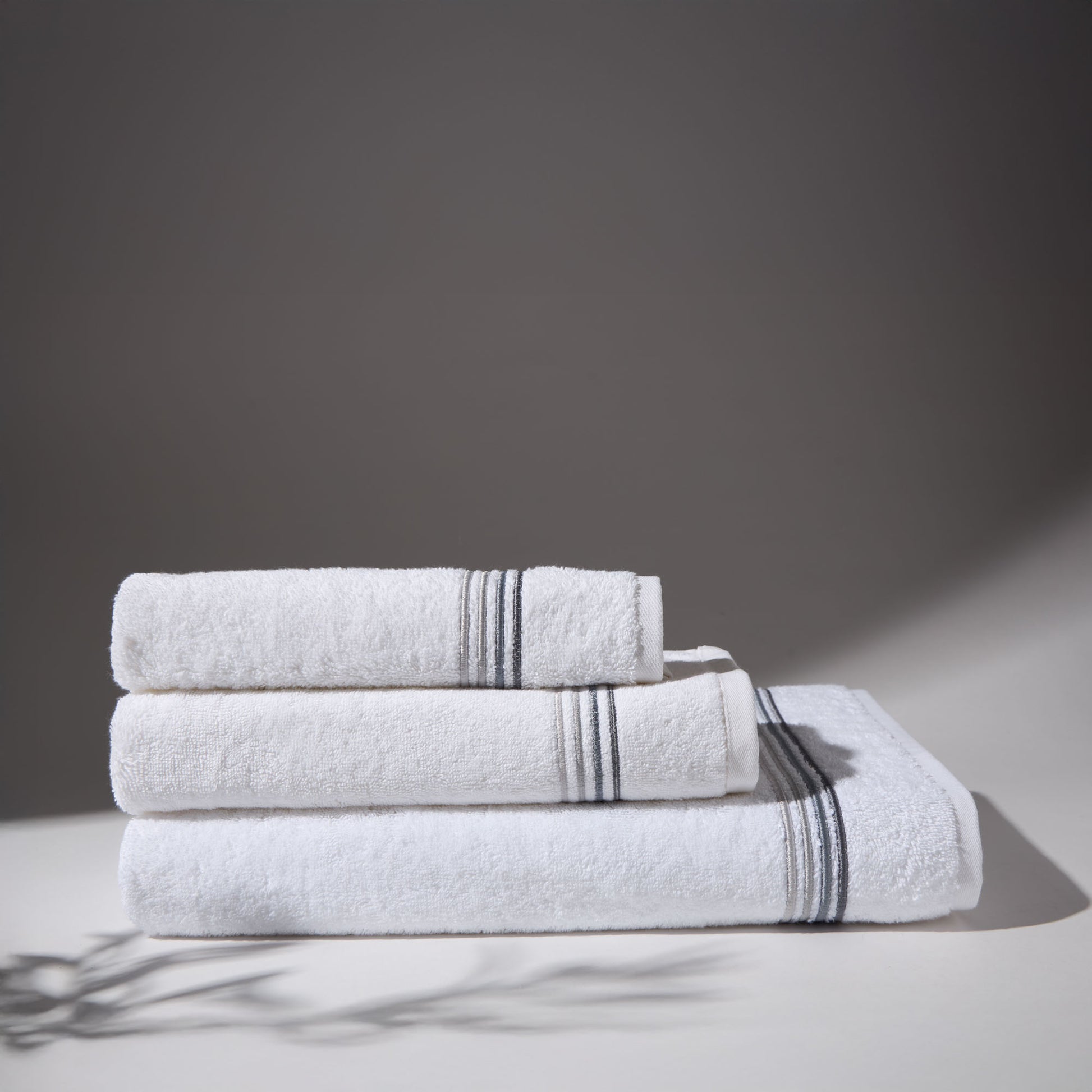Set of three white towels with gray stripes folded on a surface with a gray background.