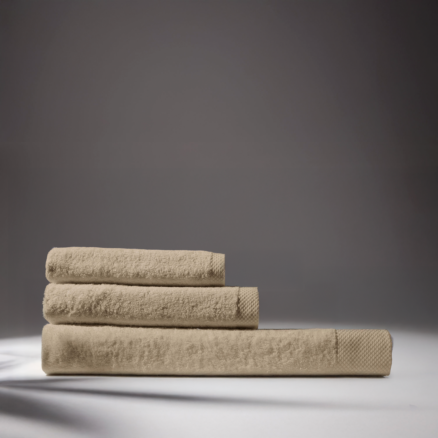 Stack of beige bath towels on a white surface, creating a minimalist and comfortable aesthetic composition, with a neutral gray background.