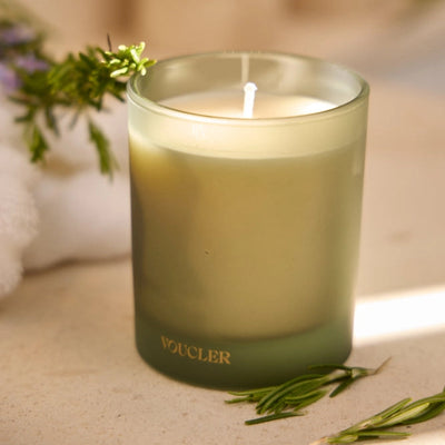 Light green scented candle with a frosted glass container and a lit flame, decorated with rosemary and mint sprigs around it, creating a relaxing atmosphere.