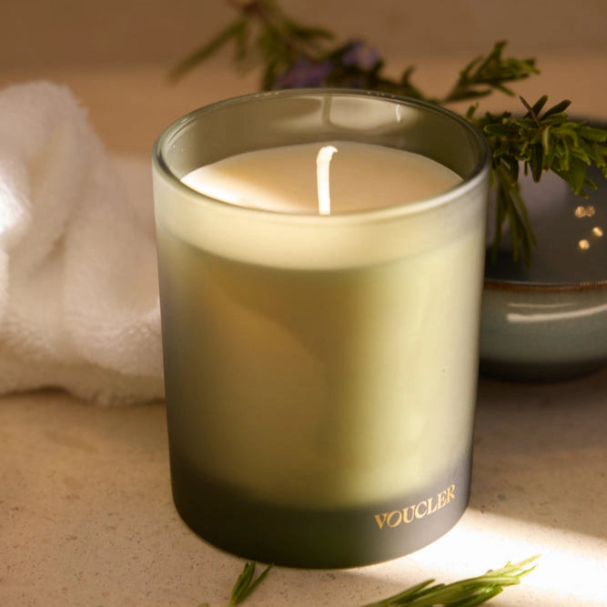 A white scented candle lit in a cozy setting, decorated with plants and a white tablecloth in the background, creating a relaxing and elegant atmosphere.