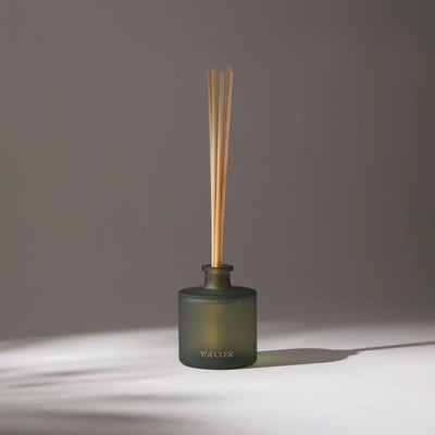 Vivid Sky aroma diffuser with wooden reeds in a dark glass container on a light surface, softly lit with soft shadows in the background.