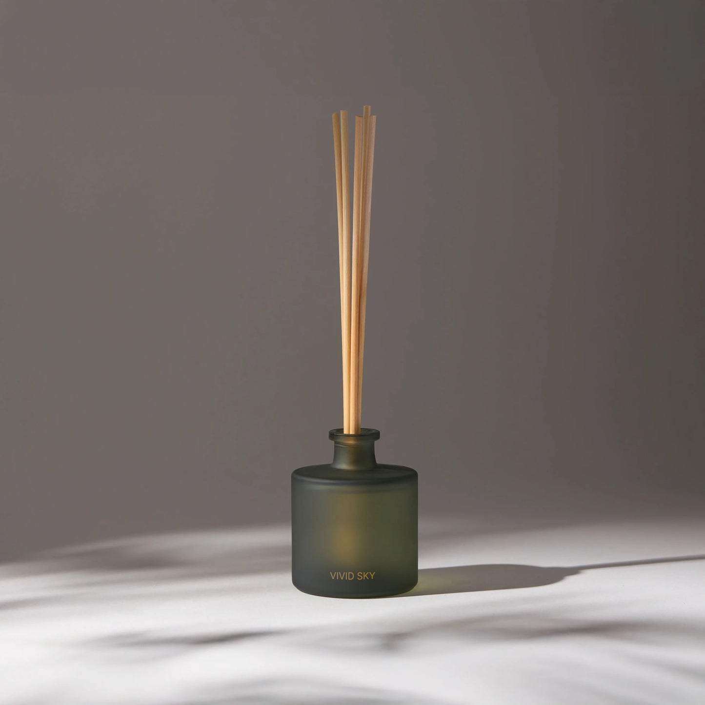 Vivid Sky aroma diffuser with wooden reeds in a dark glass container on a light surface, softly lit with soft shadows in the background.
