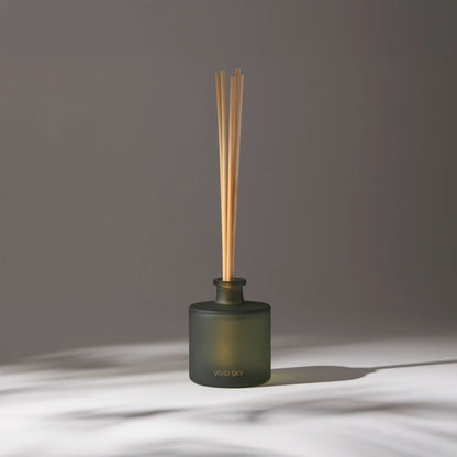 Vivid Sky aroma diffuser with wooden reeds in a dark glass container on a light surface, softly lit with soft shadows in the background.